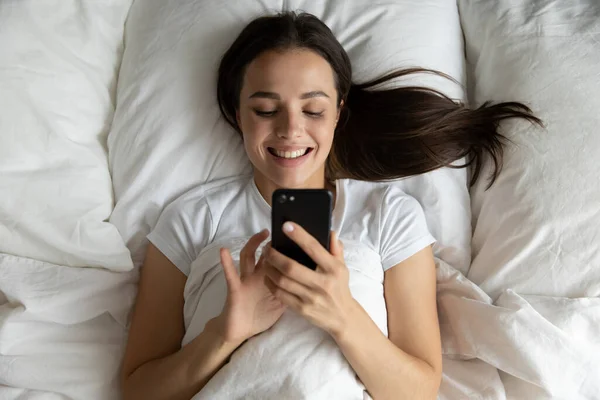 Vista superior sonriente mujer joven usando el teléfono, relajarse en la cama — Foto de Stock