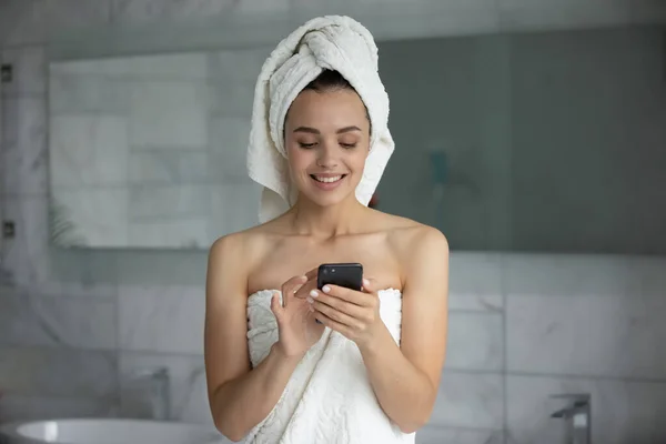 Улыбающаяся молодая женщина с помощью смартфона после душа в ванной комнате — стоковое фото