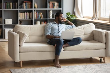 Düşünceli Afrikalı Amerikalı bir adam dizüstü bilgisayarla kanepede dinleniyor.