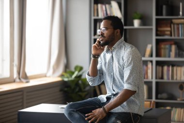 Serious African American man talking on phone, sitting on desk clipart