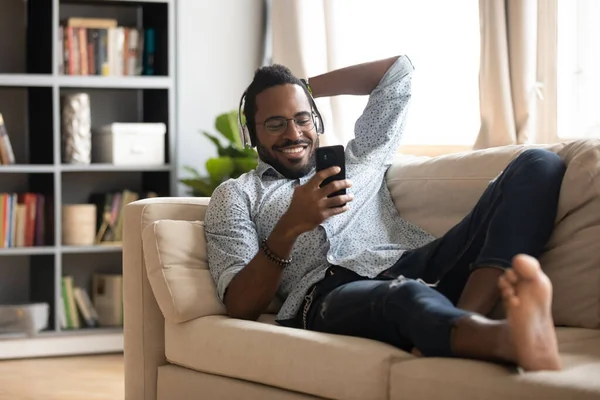 Hombre afroamericano feliz usando auriculares usando el teléfono en el sofá —  Fotos de Stock