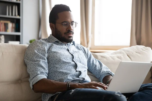 Sério focado afro-americano homem trabalhando no laptop no sofá — Fotografia de Stock