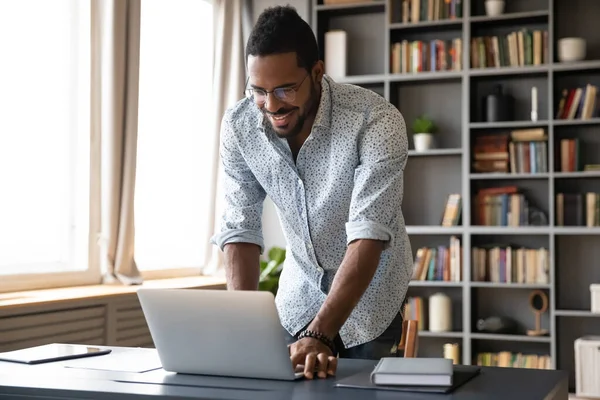 Smiling African American businessman standing near desk, using laptop — Stock Photo, Image
