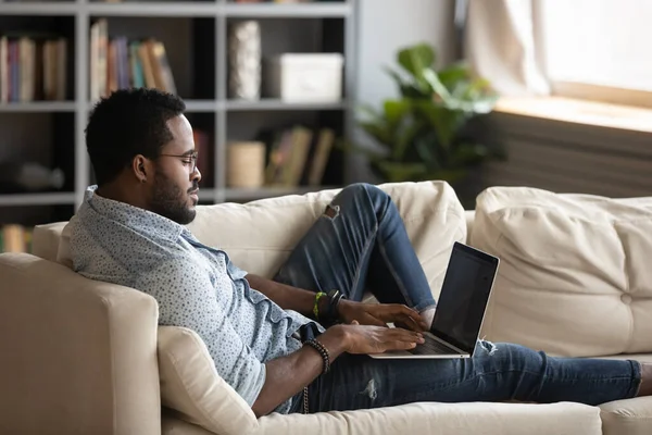 Kendine güvenen Afro-Amerikan bir adam kanepeye uzanmış, dizüstü bilgisayar kullanıyor. — Stok fotoğraf