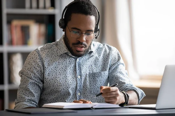 Serious focused African American man wearing headphones writing notes — Stock Photo, Image
