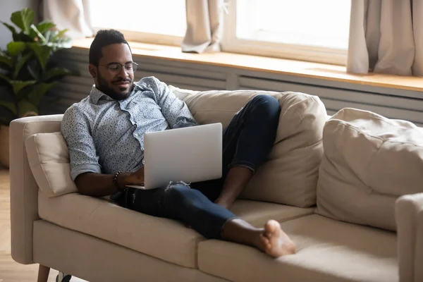 Nöjd afroamerikansk man avkopplande på soffan, med hjälp av laptop — Stockfoto