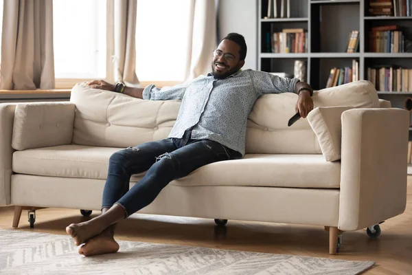 Sorrindo afro-americano homem relaxante no sofá, assistindo programa de tv — Fotografia de Stock