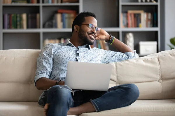 Positiv drömmande afroamerikansk man avkopplande på soffan med laptop — Stockfoto