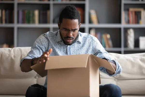 Mutsuz afro-amerikan bir adam paketini açıyor, kutunun içine bakıyor — Stok fotoğraf