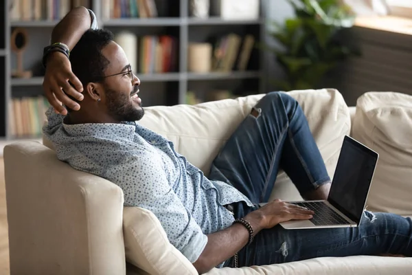 Drömmande leende afroamerikansk man liggande på soffan med laptop — Stockfoto