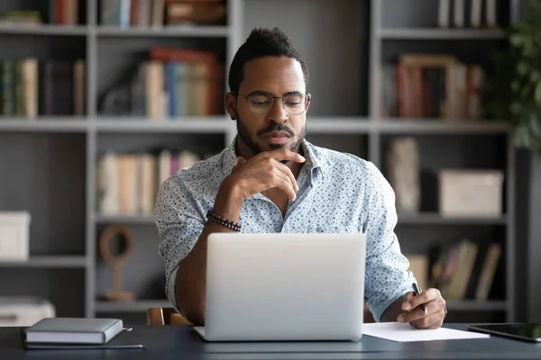 Thoughtful African American businessman looking at laptop screen, touching chin — Stock Photo, Image