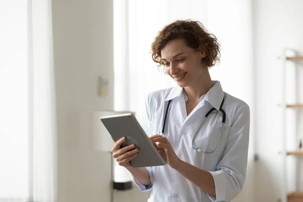 Smiling woman professional doctor using digital tablet, standing in office