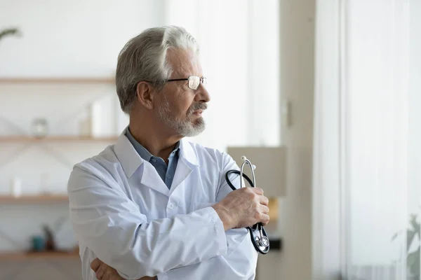 Bedachtzame volwassen arts draagt bril en uniform verloren in gedachten — Stockfoto