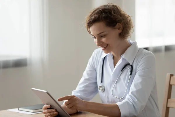 Sonriente joven doctora usando tableta digital, sentada en el escritorio —  Fotos de Stock