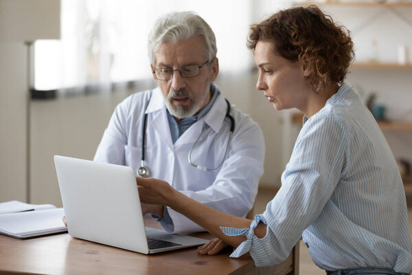 Serious mature doctor showing checkup result on laptop to patient