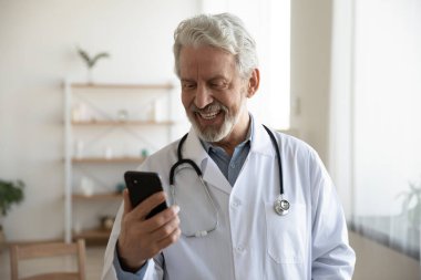 Beyaz üniformalı gülümseyen olgun doktor akıllı telefon ekranına bakıyor.