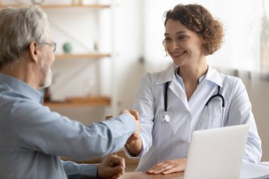 Gülümseyen genç doktor toplantıda olgun hasta elini sıkıyor.