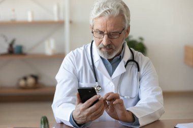 Ciddi bir yetişkin doktor akıllı telefon ekranına bakıyor, masada oturuyor.