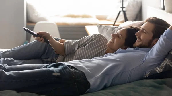 Glada avslappnade par liggande på sängen, med hjälp av fjärrstyrd tv. — Stockfoto
