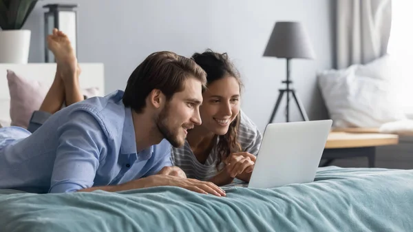 興味のある若いカップルがベッドの上に横たわって、コンピュータ画面を見て. — ストック写真