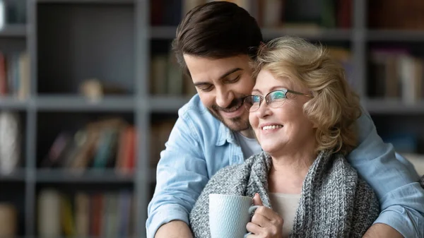 Lächelnder junger Mann umarmt reife Mutter, die Tee trinkt — Stockfoto