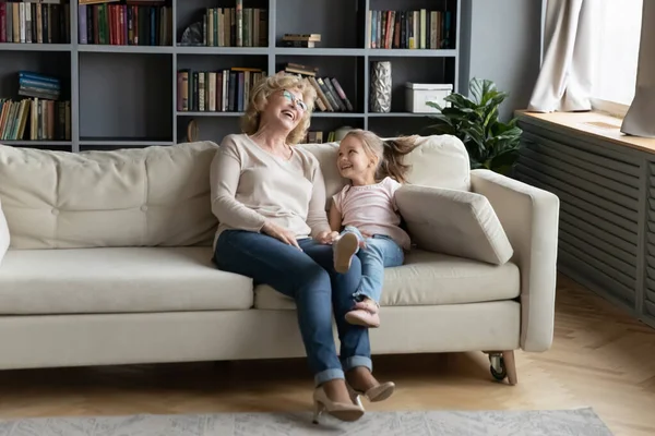Веселая маленькая внучка и зрелая бабушка смеются, сидя на диване — стоковое фото