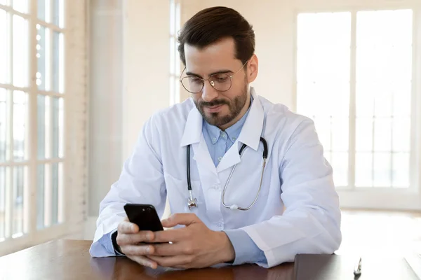 Zaostřený lékař nosí brýle pomocí smartphonu, sedí za pracovním stolem — Stock fotografie