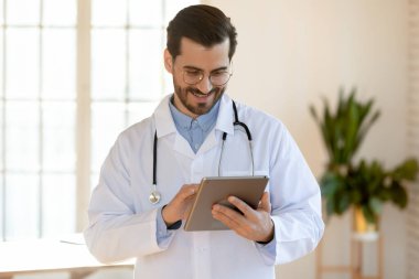 Gülümseyen doktor gözlük takıyor, tablet kullanıyor, ofiste duruyor.