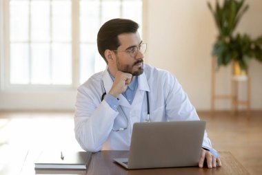 Gözlük takan düşünceli bir doktor dizüstü bilgisayarda çalışıyor, problem çözümü üzerinde kafa yoruyor.