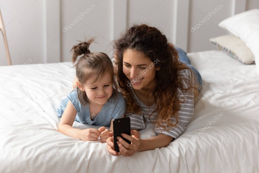 Mother lying on bed with daughter having fun using smartphone