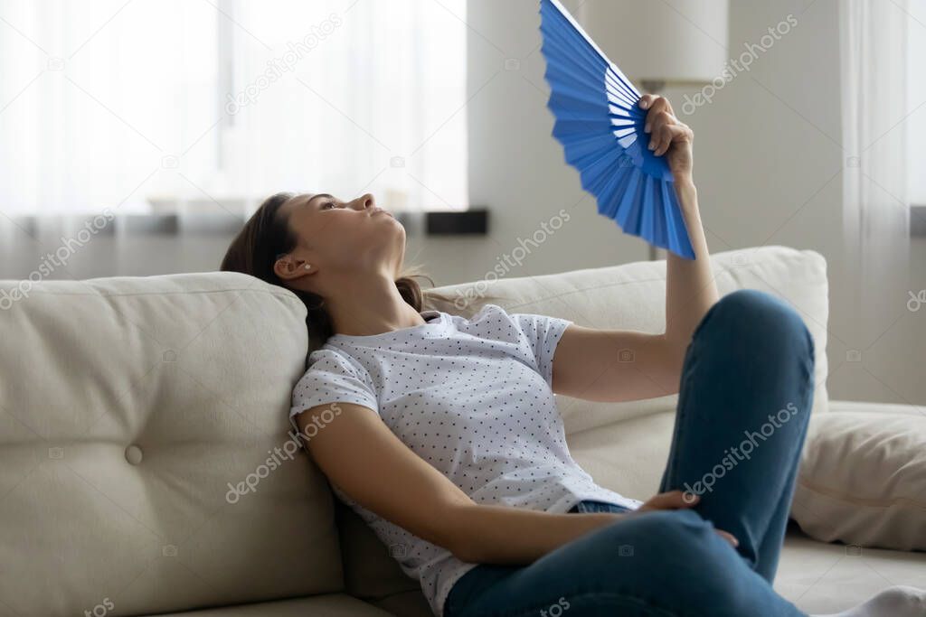 Exhausted young woman waving paper fan, suffering from high temperature.
