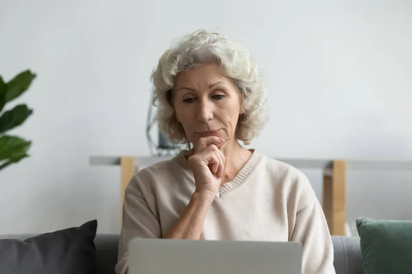 Ernste Frau mittleren Alters blickt auf Laptop-Bildschirm, berührt Kinn — Stockfoto