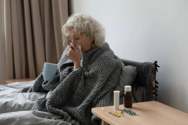 Kranke reife Frau bläst laufende Nase, sitzt im Bett — Stockfoto