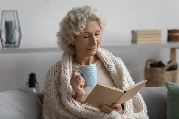 Friedliche zufriedene reife Frau in Decke gehüllt Lesebuch zu Hause — Stockfoto