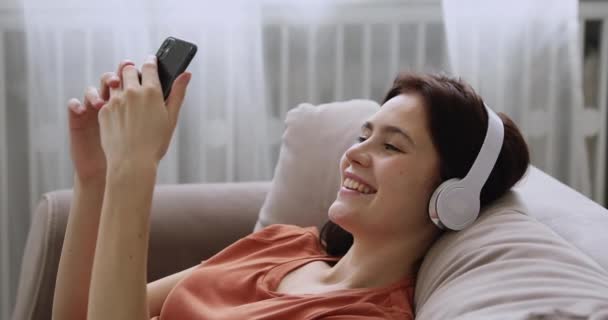女孩头戴耳机，手持智能手机靠在沙发上，在室内放松 — 图库视频影像