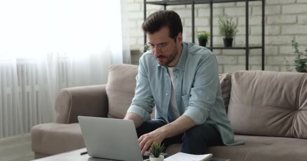 Ο άνθρωπος κάθεται στον καναπέ χρήση laptop διαβάστε ειδήσεις αισθάνεται χαρούμενος — Αρχείο Βίντεο