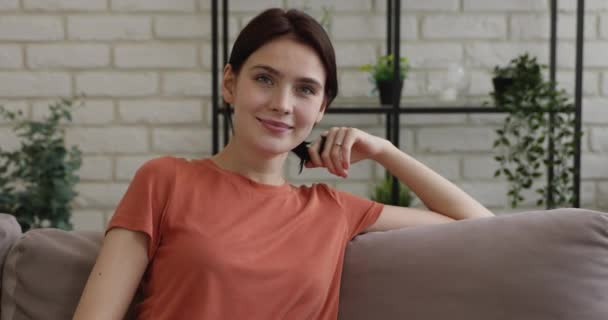 Chica sentada en el sofá en casa sonriendo mirando a la cámara — Vídeos de Stock