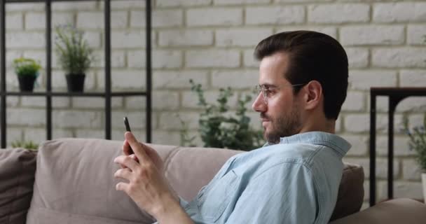 Guy απολαμβάνουν ελεύθερο χρόνο κάθονται ακουμπισμένα στον καναπέ με smartphone — Αρχείο Βίντεο
