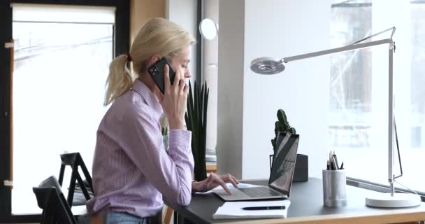 Mujer de negocios sentarse en el escritorio de trabajo con el ordenador portátil hablando en el teléfono inteligente — Vídeos de Stock