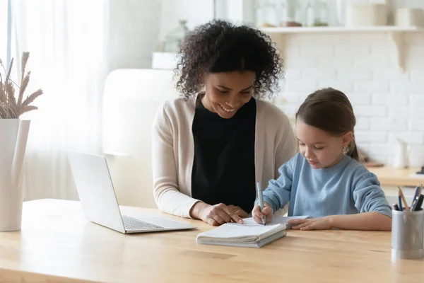 Sorridente misto razza madre aiutare poco adottato figlia con compiti a casa. — Foto Stock