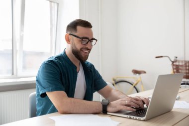 Gülümseyen iş adamı gözlük takıyor, dizüstü bilgisayar kullanıyor, masada oturuyor.