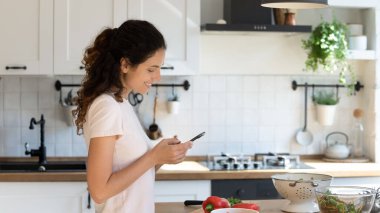 Evde cep telefonuyla yemek pişiren mutlu genç bir kadın.