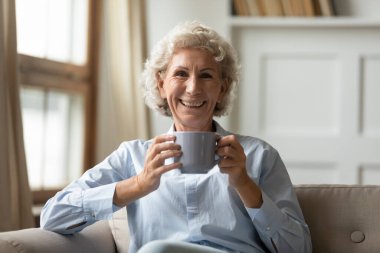 Güzel, zarif, yaşlı bir kadın elinde bir bardak içkiyle evin içinde poz veriyor.