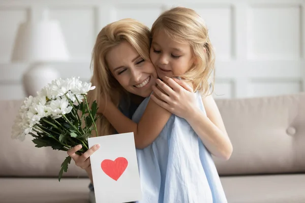 Millennial matka obejmująca małą dziewczynkę trzymającą kwiaty i kartkę z życzeniami — Zdjęcie stockowe