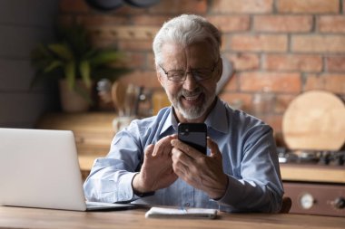 Gülümseyen yaşlı adam bilgisayarında akıllı telefon kullanıyor.
