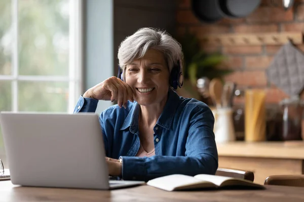 Smart elderly woman in headphones watch webinar on laptop