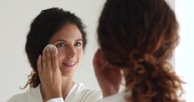 Aynada yansıyan güzel bir kadın elinde pamuk tutarak deriyi temizler.