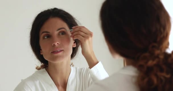 Beautiful woman looking in mirror holding tweezers plucks eyebrows — Stock Video