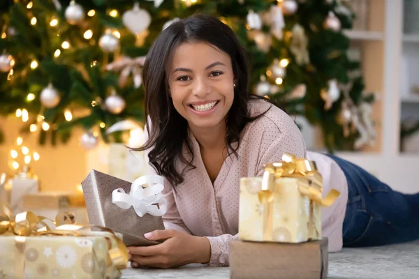 Mujer biracial sonriente tendida cerca del árbol de Navidad con regalos envueltos — Foto de Stock