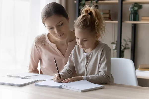 Cuidar a la joven madre ayudando a su pequeña hija con la tarea escolar —  Fotos de Stock